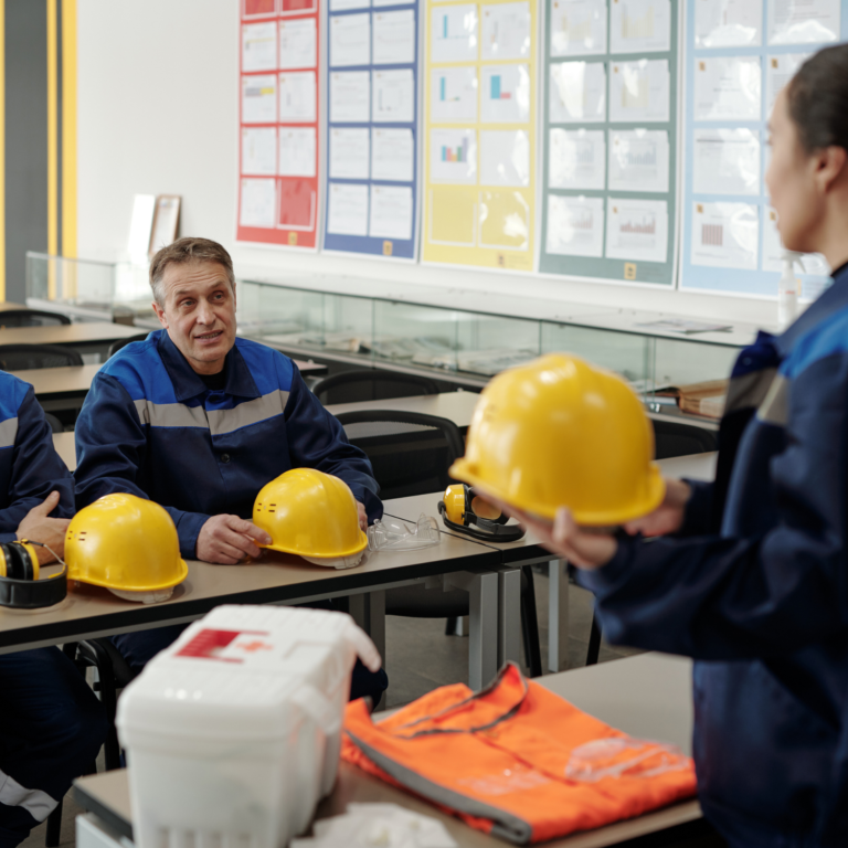 Grupo de trabajadores en una sala de formación, escuchando a una instructora con cascos amarillos.
