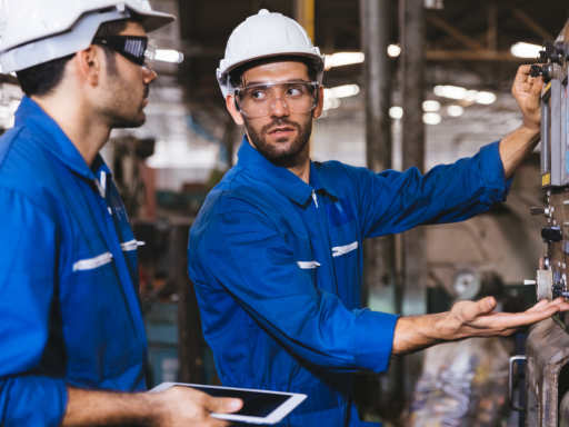 Dos trabajadores en un entorno industrial, uno usando una tablet y ambos con cascos y gafas.