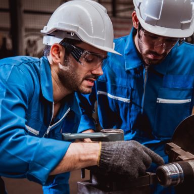 Dos trabajadores en overol azul, usando cascos, revisando maquinaria en un taller.